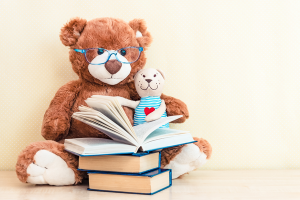 Nounours en peluche avec des lunettes entrain de lire un livre ouvert, posé sur une pile de livres.
