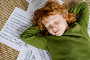 Photo d'un enfant au cheveux roux et avec un pull faire allongé les yeux fermés sur des partitions répendues au sol